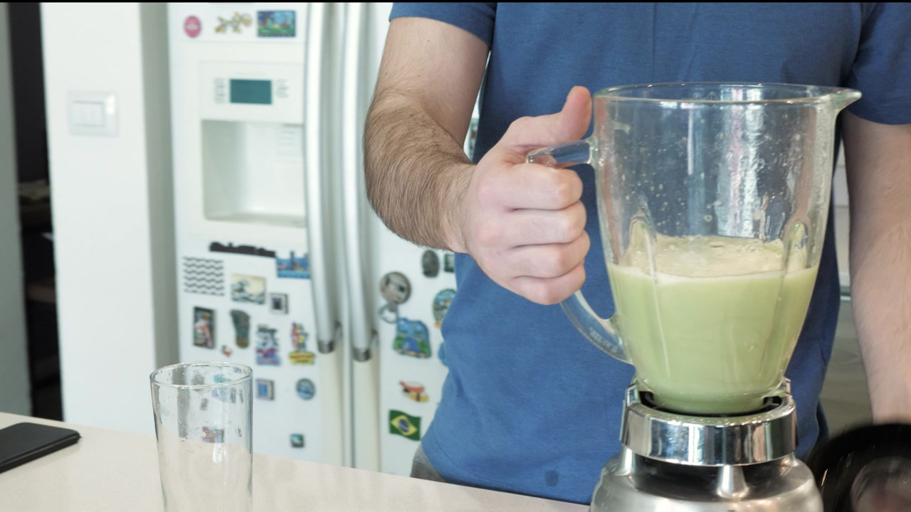 A bearded man wearing a blue t-shirt prepares a green juice in a blender on a kitchen counter, serves the juice in a glass and takes a sip, in the background a refrigerator covered in magnets, a kitchen sink and a large window