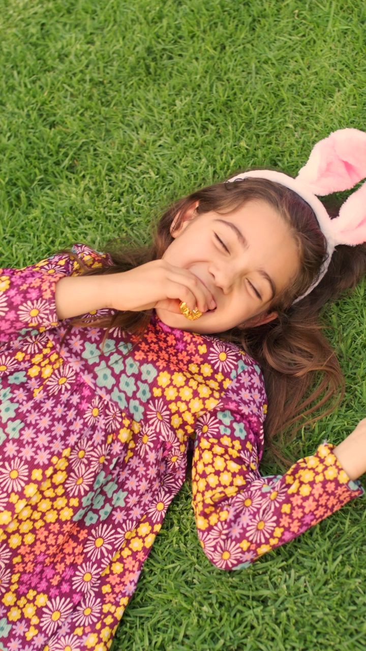 A birds eye view shot of a little girl laying in the grass while eating a chocolate bunnie, a little girl wearing a flower pattern dresss smiles to the camera while eating a chocolate