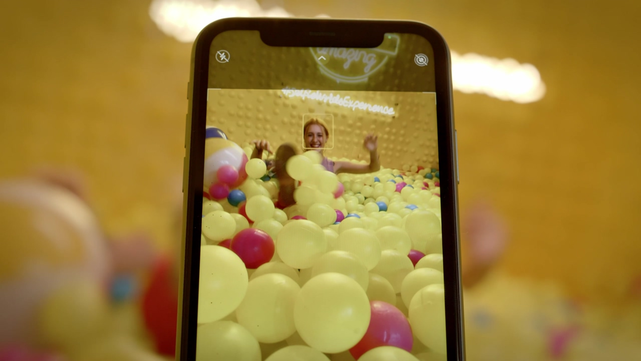 A black smartphone records a young happy woman playing in the plastic ball pit pool for a social media video