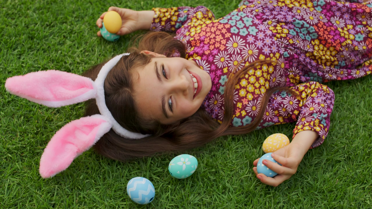 A girl wearing white bunny ears is lying on the grass with decorated easter eggs as she smiles and looks up at the sky
