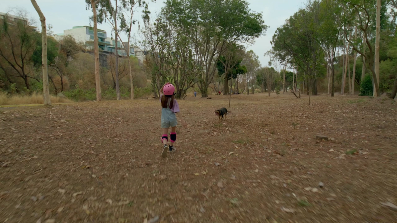 A little girl wearing a pink helmet and denim overall runs through the forest to find her little brown dog and then she pets him on the back