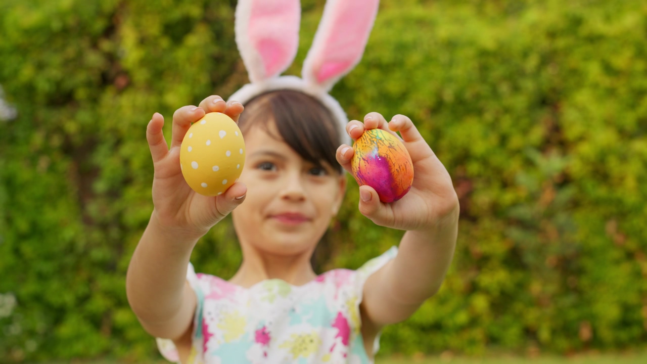A little girl wearing bunny ears shows decorated easter eggs to the camera