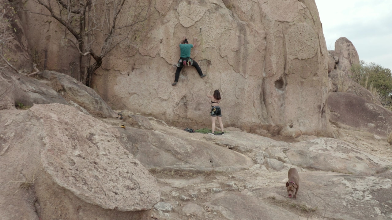 A man and a woman climbers starting to climb a gigantic rock outside, with the help of ropes and harnesses