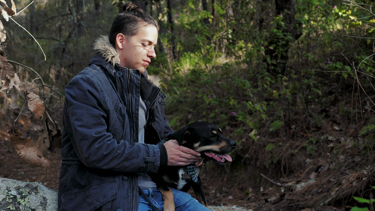 A man wearing a blue hooded jacket sits on a rock petting a black and white dog next to him, trees in the background