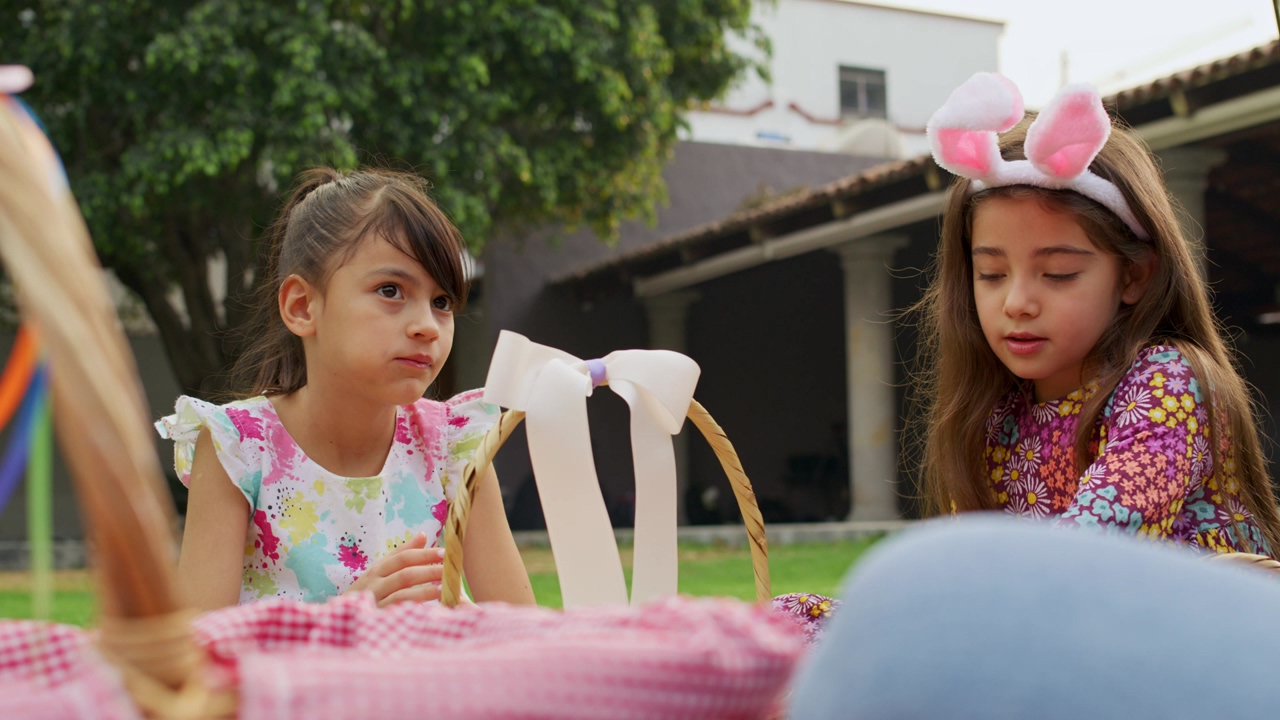 A pair of little girls eating rabbit shaped chocolates on an easter day next to baskets full of easter eggs