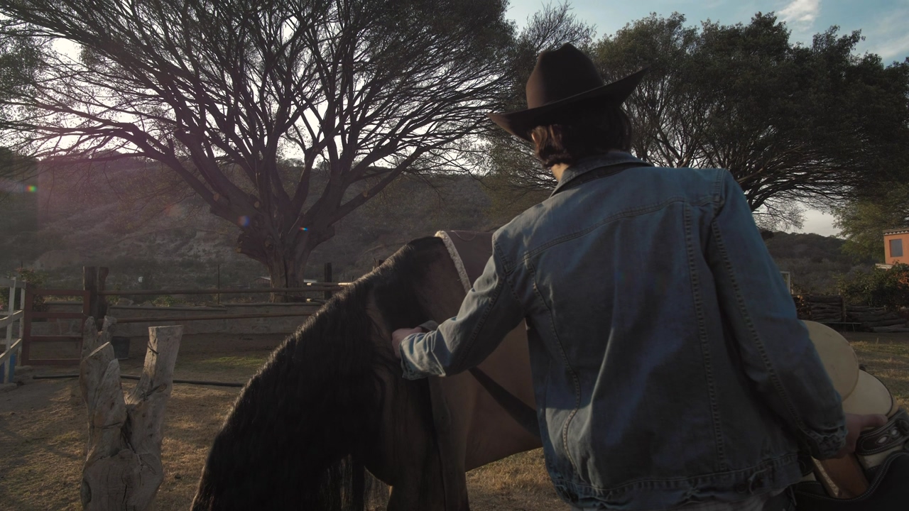 A person wearing a black cowboy hat, a denim jacket and blue jeans puts a saddle on a brown horse with a black mane, trees in the background