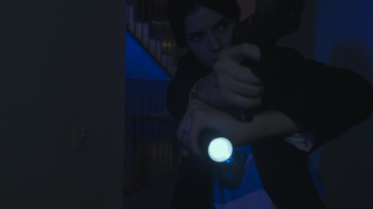 A policewoman enters a dark apartment pointing a gun and flashlight straight ahead, moving slowly through the room, with patrols outside with their turrets on