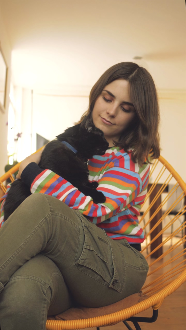 A woman wearing a colorful striped sweater caresses a black cat with a blue collar while sitting in a yellow chair at home