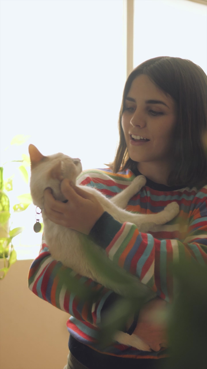 A woman wearing a colorful striped sweater caresses and talks with a white cat at home