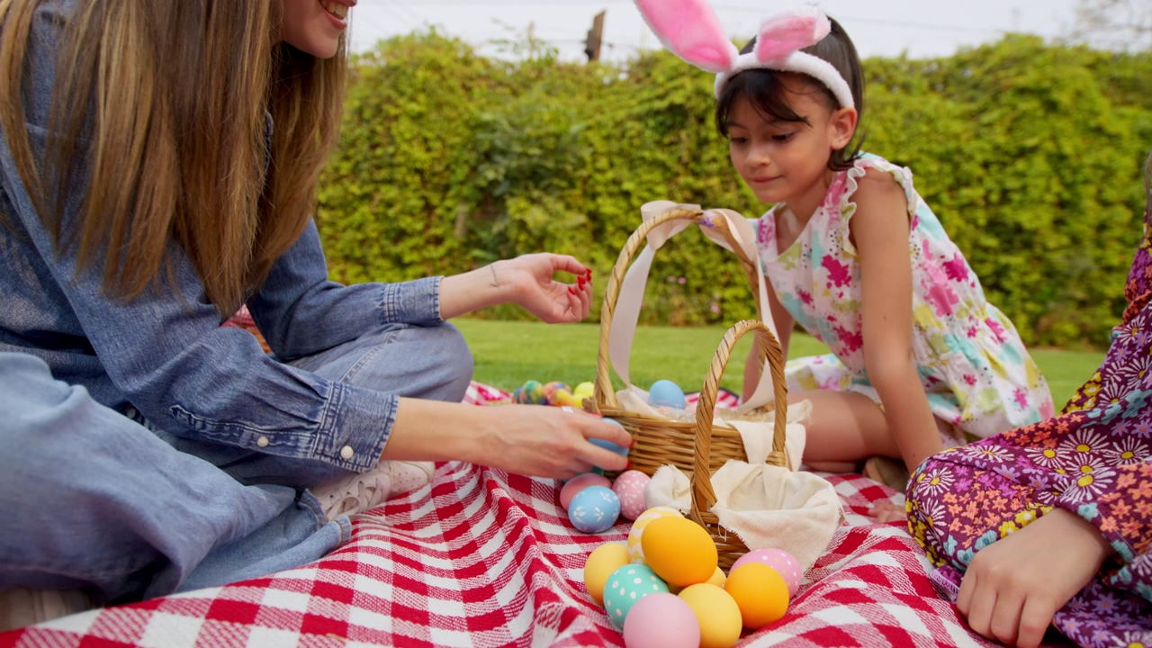 A young woman is teaching two little girls how to separate easter eggs in the backyard garden, two little girls with easter egg bunny costumes watch a woman separates the easter eggs in the basket
