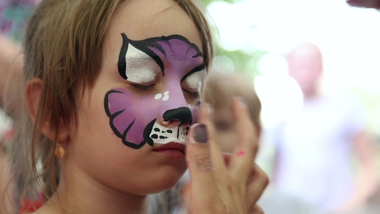 Adding glitter to face paint of a purple cat on a child, child, artist, cat, makeup artist, glitter, and face paint