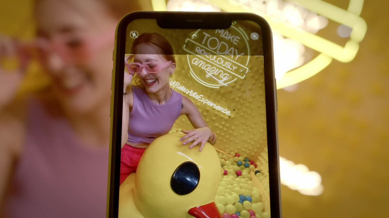 Close up shot of a black smartphone recording a young influencer having fun riding a giant rubber duck in a plastic ball pool with a neon sign in the background