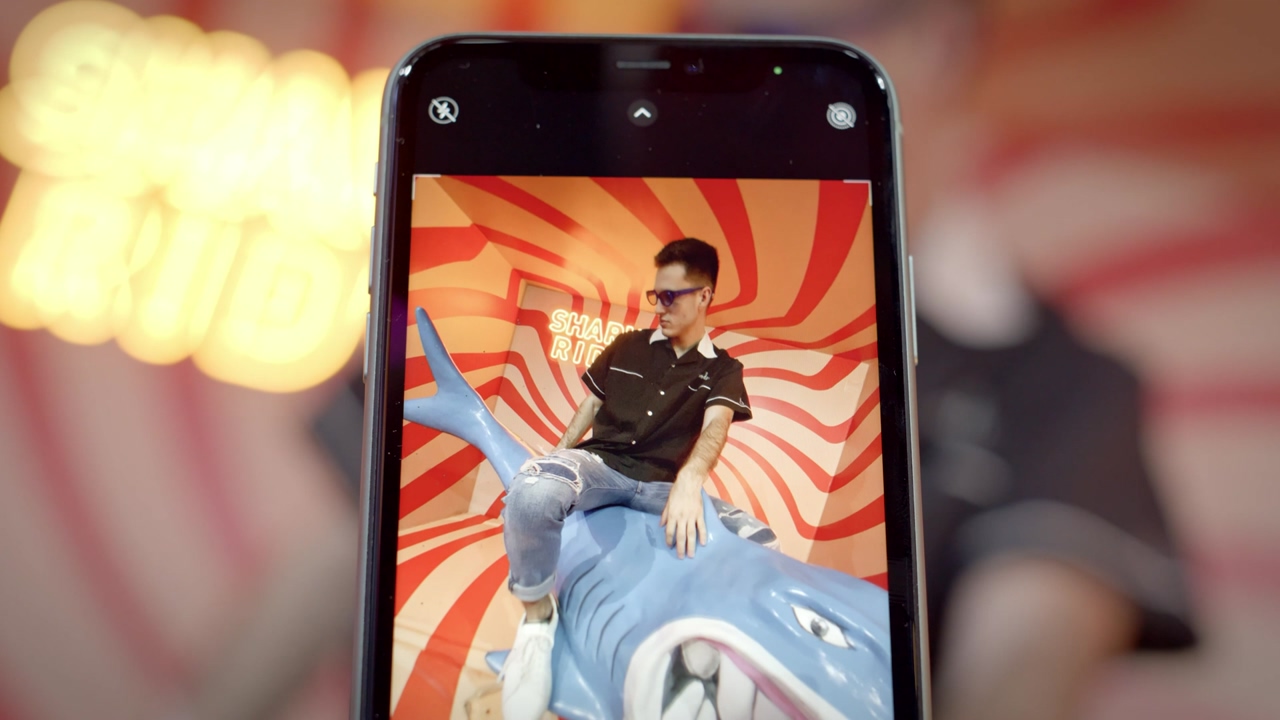 Close up shot of a black smartphone recording a young man with sunglasses riding a fake shark for social media posts in an orange room with a wavy pattern
