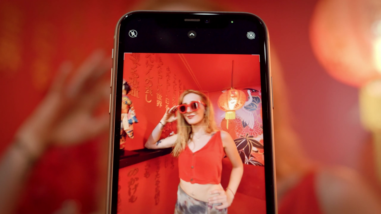 Close up shot of a smartphone recording a young influencer woman wearing red sunglasses posing and giggling in a china-themed room for a social media video