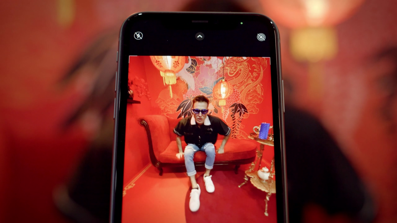 Close up shot of a smartphone recording a young urban a young man wearing sunglasses in a red china-themed room posing on a red couch for the smarphone