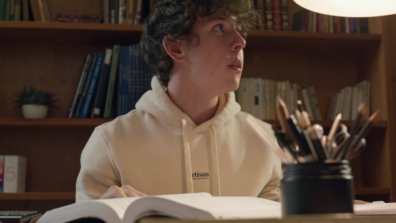 Close up shot of a young man wearing a hoodie that dedicates his evening to studying in the library, accompanied by a trusty calculator and the warm of the desk lamp