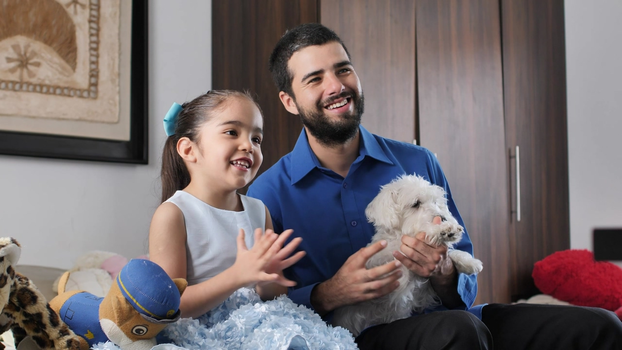 Father and daughter sitting in a room surrounded by stuffed animals while watching tv and reacting together with laughter, the father has a small white dog on his legs