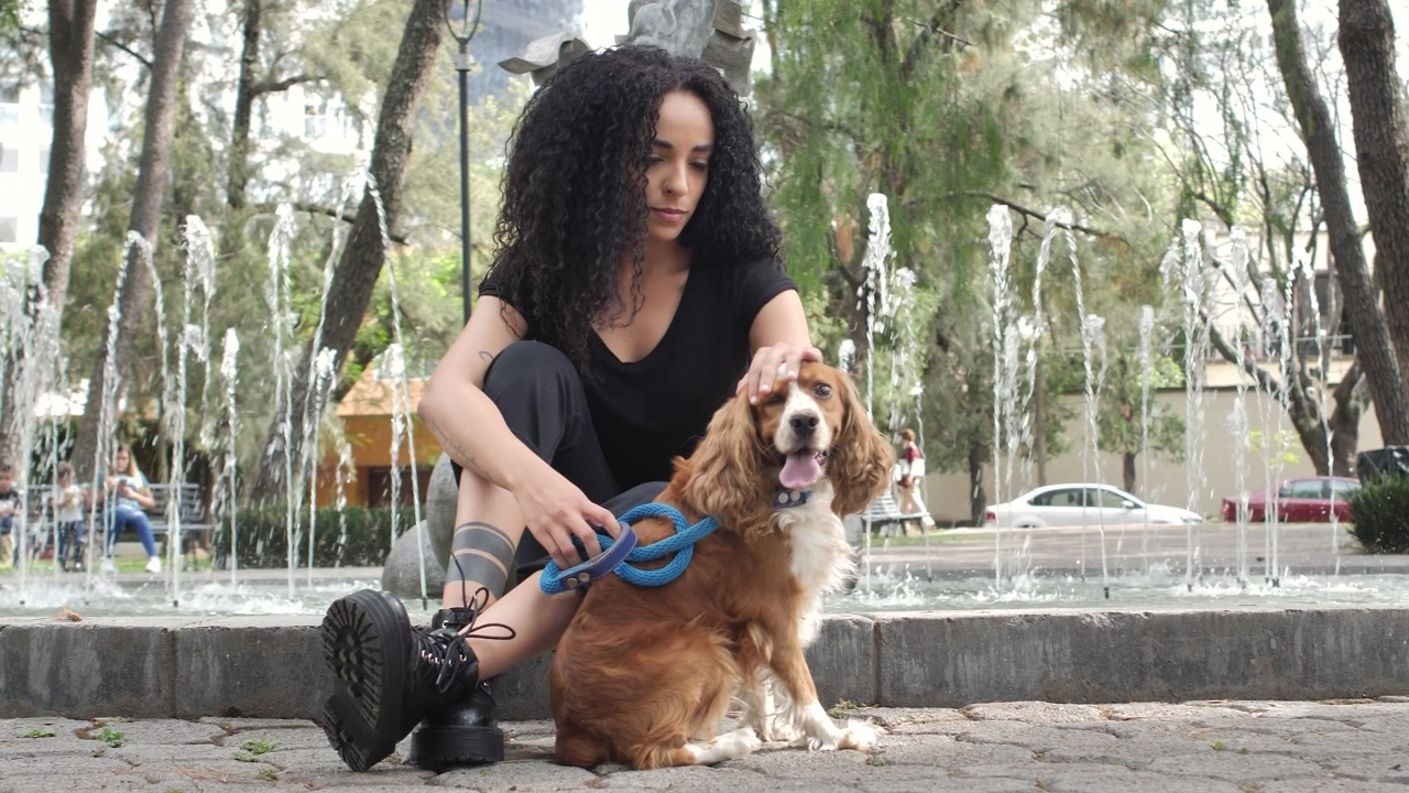 Girl sitting on the edge of a fountain in a park, with her dog at her side while lovingly petting him, and the camera pans from side to side