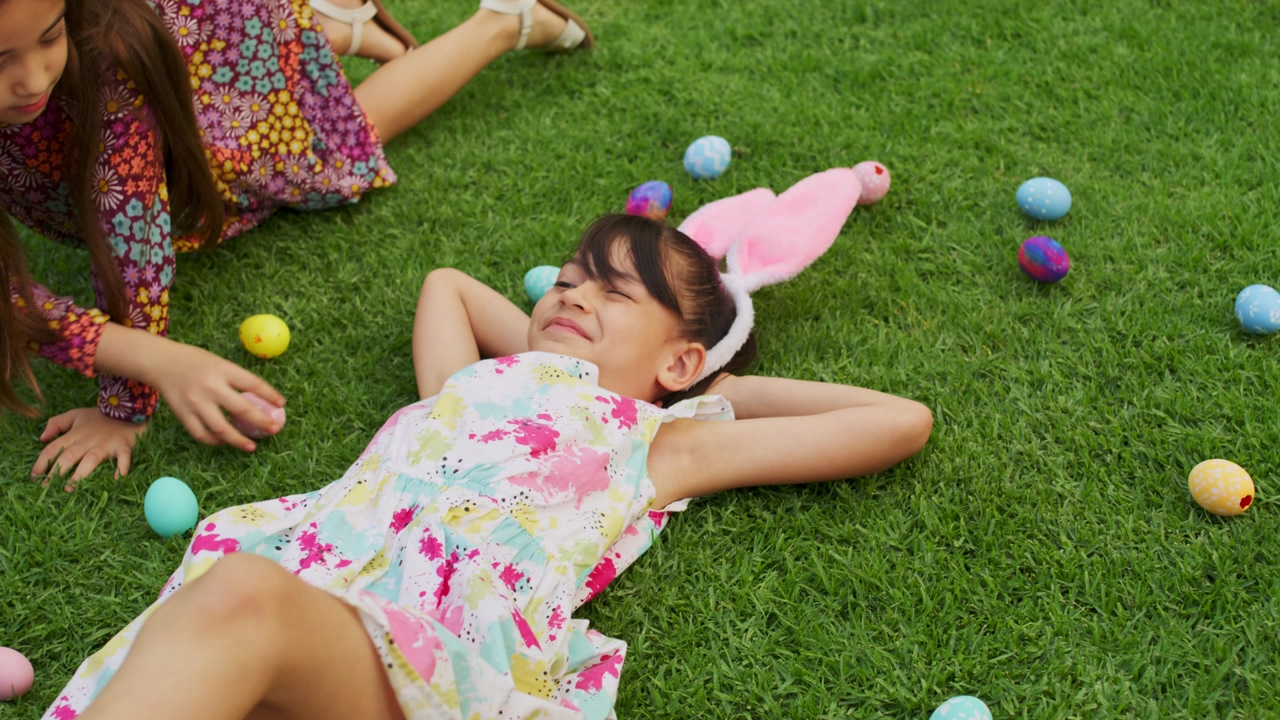 Girls play in their backyard grass surrounded by decorated easter eggs while wearing bunny ears on their heads
