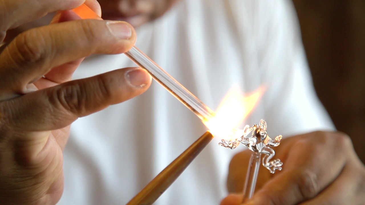 Hands of a craftsman making the figure of a glass frog near a lit blowtorch
