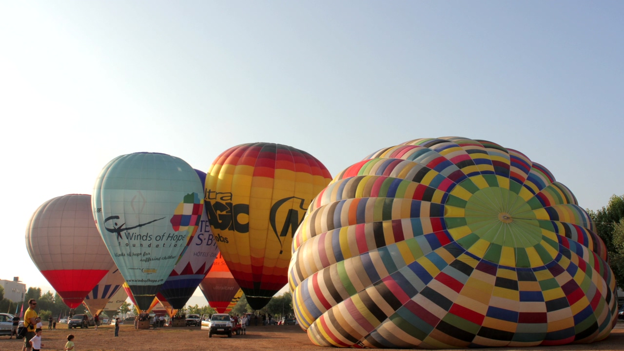 Hot air balloons of various colors take off from the ground while people see them, blue sky in the background