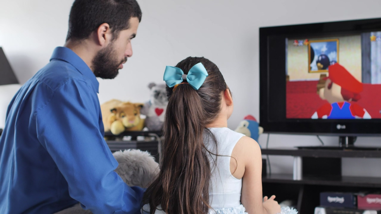 Little girl with her father seen from the back in front of a television with some stuffed animals around, playing a mario bros video game, while she explains to him how to play
