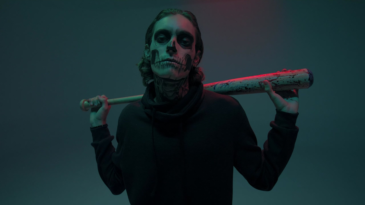 Portrait of a young man with his face painted as a skull with a bat looking at the camera, on a dark blue background