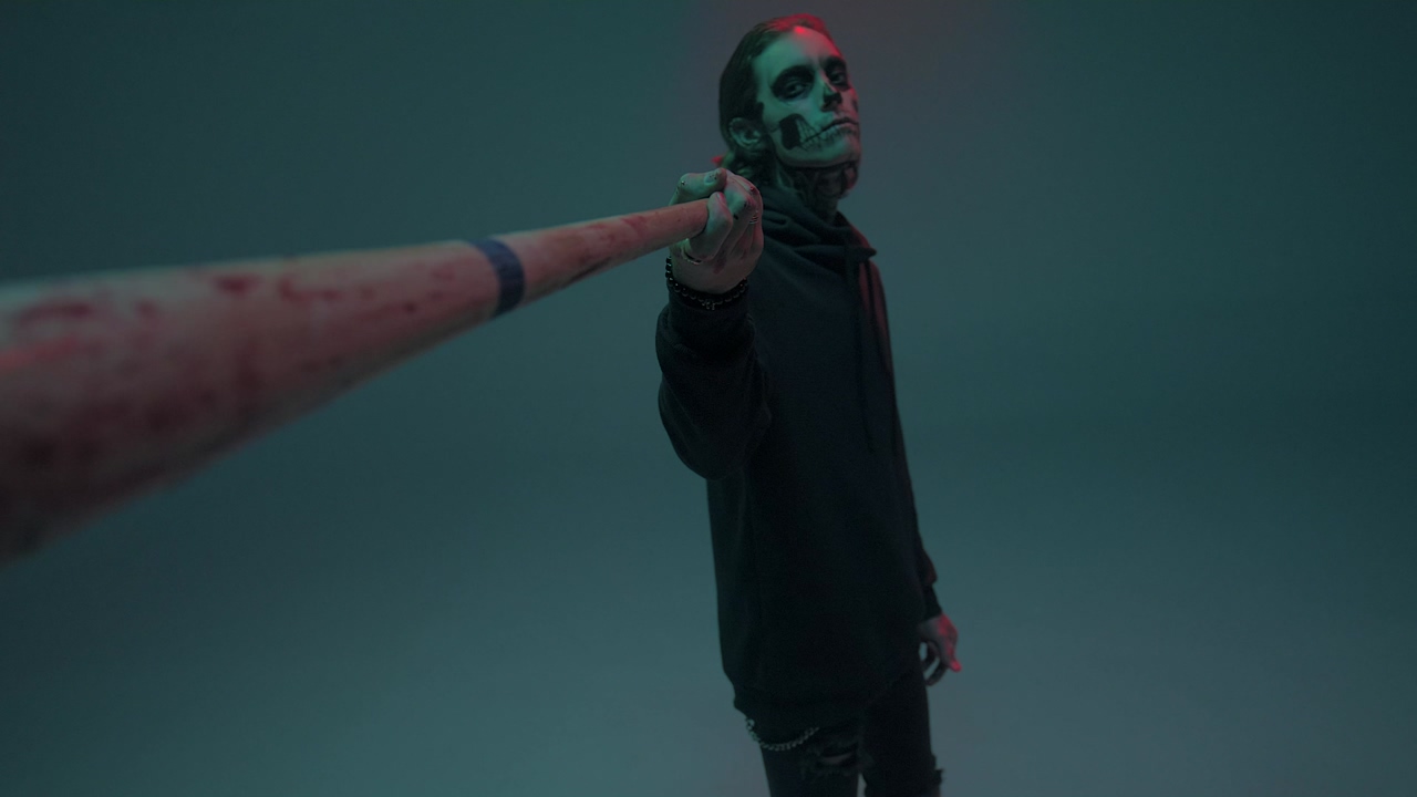 Young man dressed as a skull, pointing at the camera with a bat, on a blue background, under colored lights