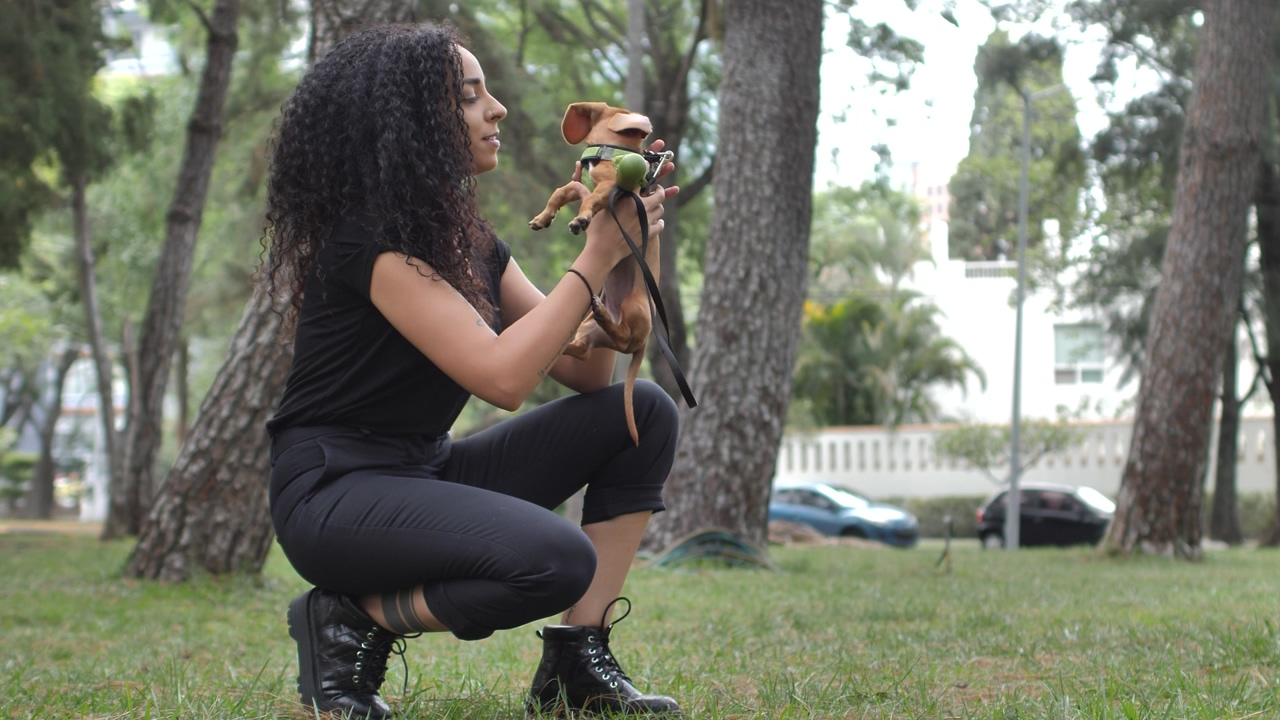 Young woman kneeling in the middle of a park with grass and trees, while she carries her little dog, and gives her tender expressions of affection