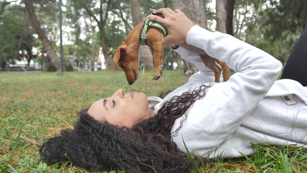 Young woman lying on the grass in the middle of a park with trees, while holding her little dog in her hands, he licks her face and she says some words to him