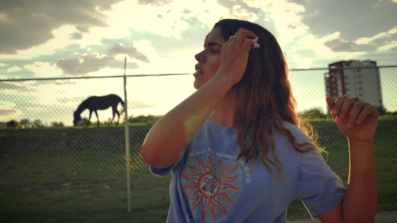 Young woman placing a flower on her ear #with a sunset behind her #a natural environment surrounded by horses #trees and a building in the distance